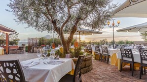 Un espace de restauration en plein air, avec des tables disposées pour deux personnes, entouré d’oliviers et offrant une vue sur le coucher du soleil.