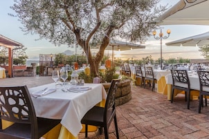Un área de comedor al aire libre con mesas preparadas para dos, rodeada de olivos y vistas a la puesta de sol.