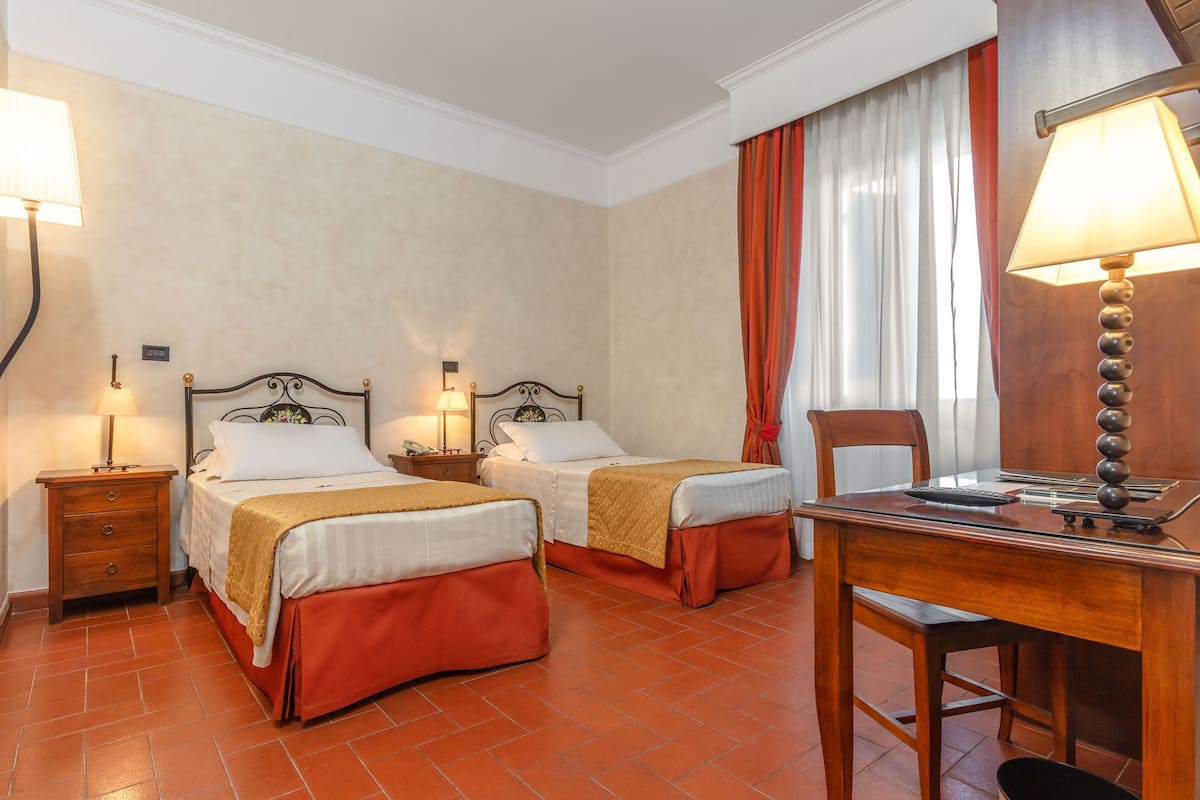 A hotel room with two beds, a desk, and a chair. There are two bedside tables with lamps.