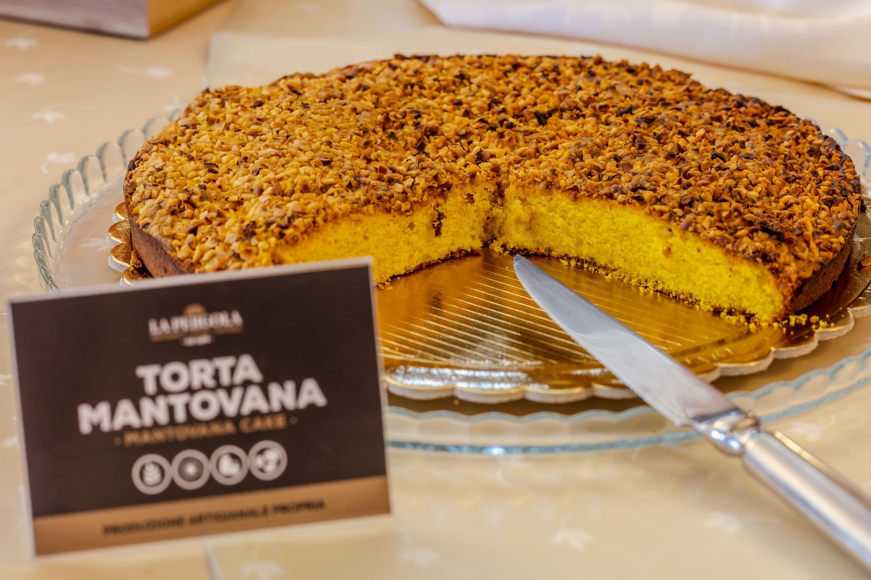 A Torta Mantovana cake with a slice cut out, placed on a glass plate with a sign beside it.