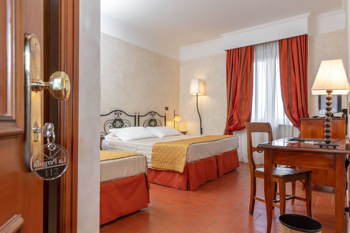 A hotel room with two beds, a desk with a lamp, a chair, and a trash bin.