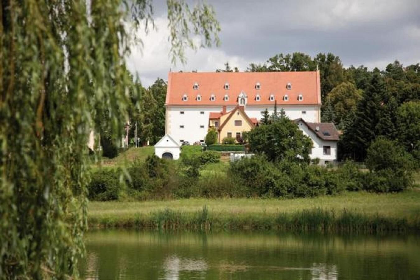 A large, white building with a red roof, surrounded by greenery and a body of water.