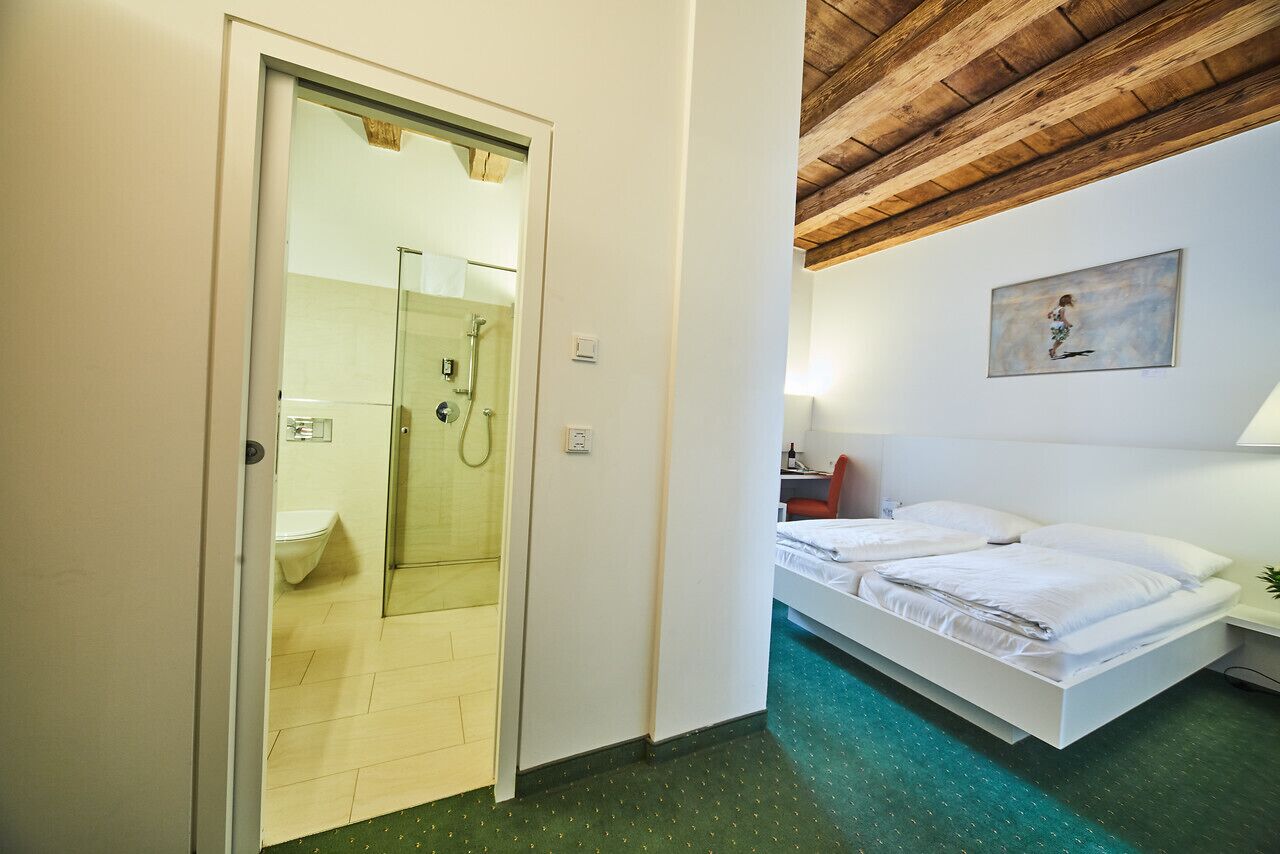 Hotellihuone, jossa on lasiseinäinen suihku, valkoisilla lakanoiden peitetty sänky ja seinällä taulu.