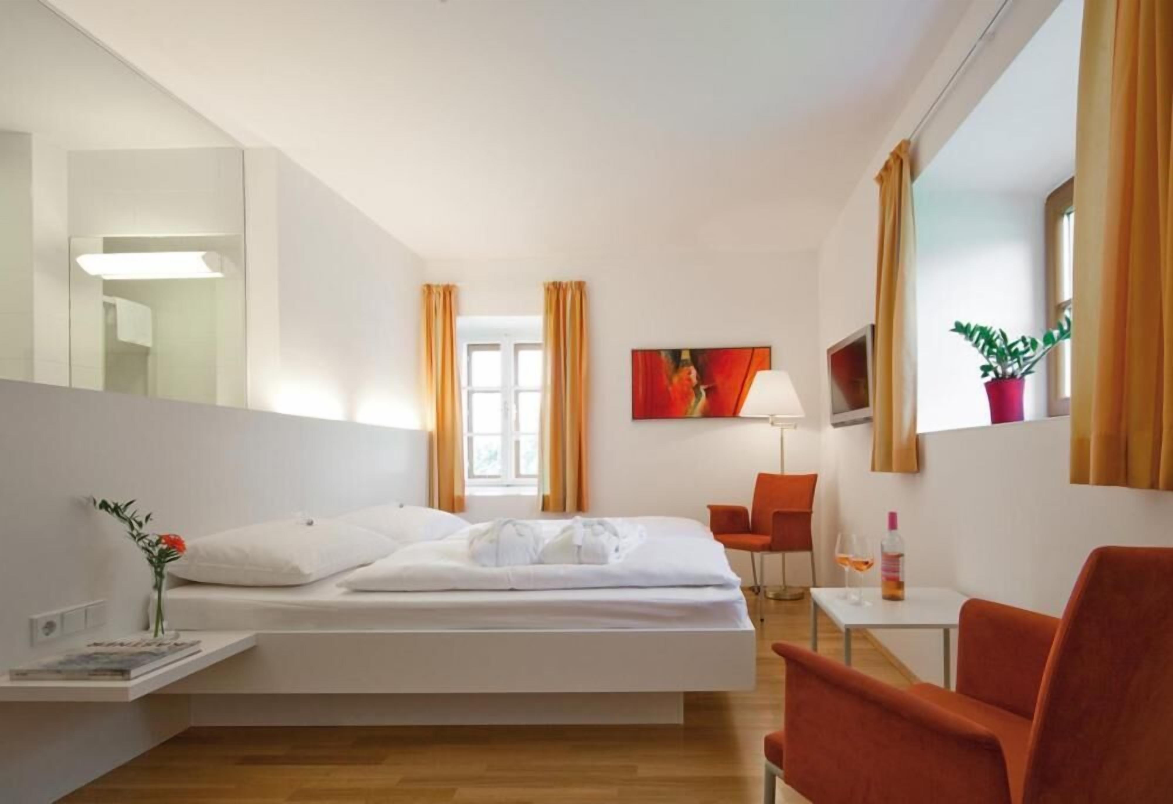 A hotel room with a bed, a chair, a table, a lamp, a painting, and a potted plant.