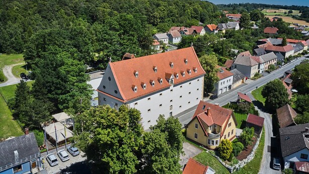 An aerial view of a town with a prominent white building with a red roof, surrounded by residential houses and lush greenery.