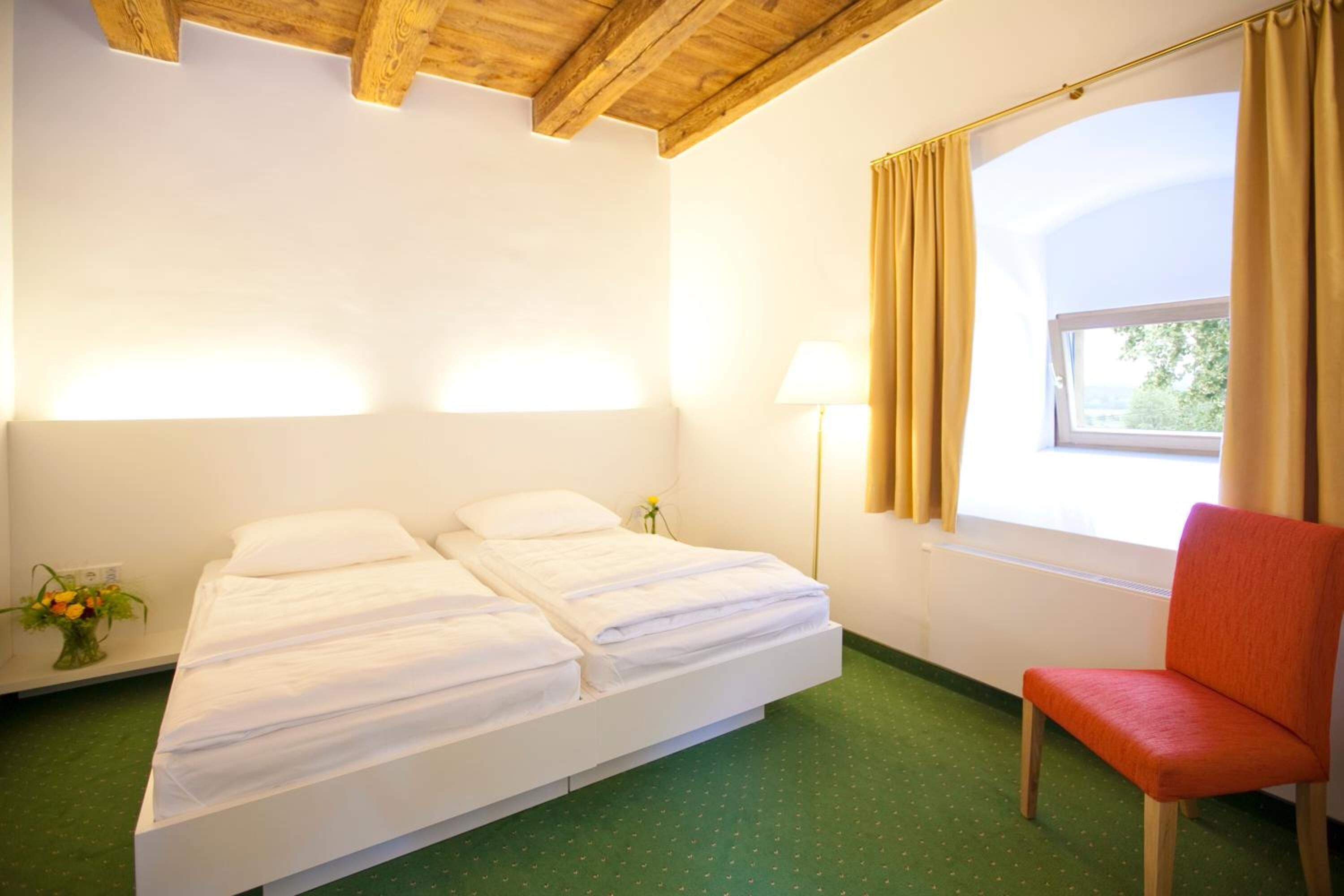 Una habitación con cama, una silla, ventana con cortinas, lámpara y un jarrón con flores.