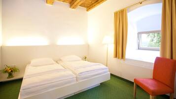 Una habitación con cama, una silla, ventana con cortinas, lámpara y un jarrón con flores.
