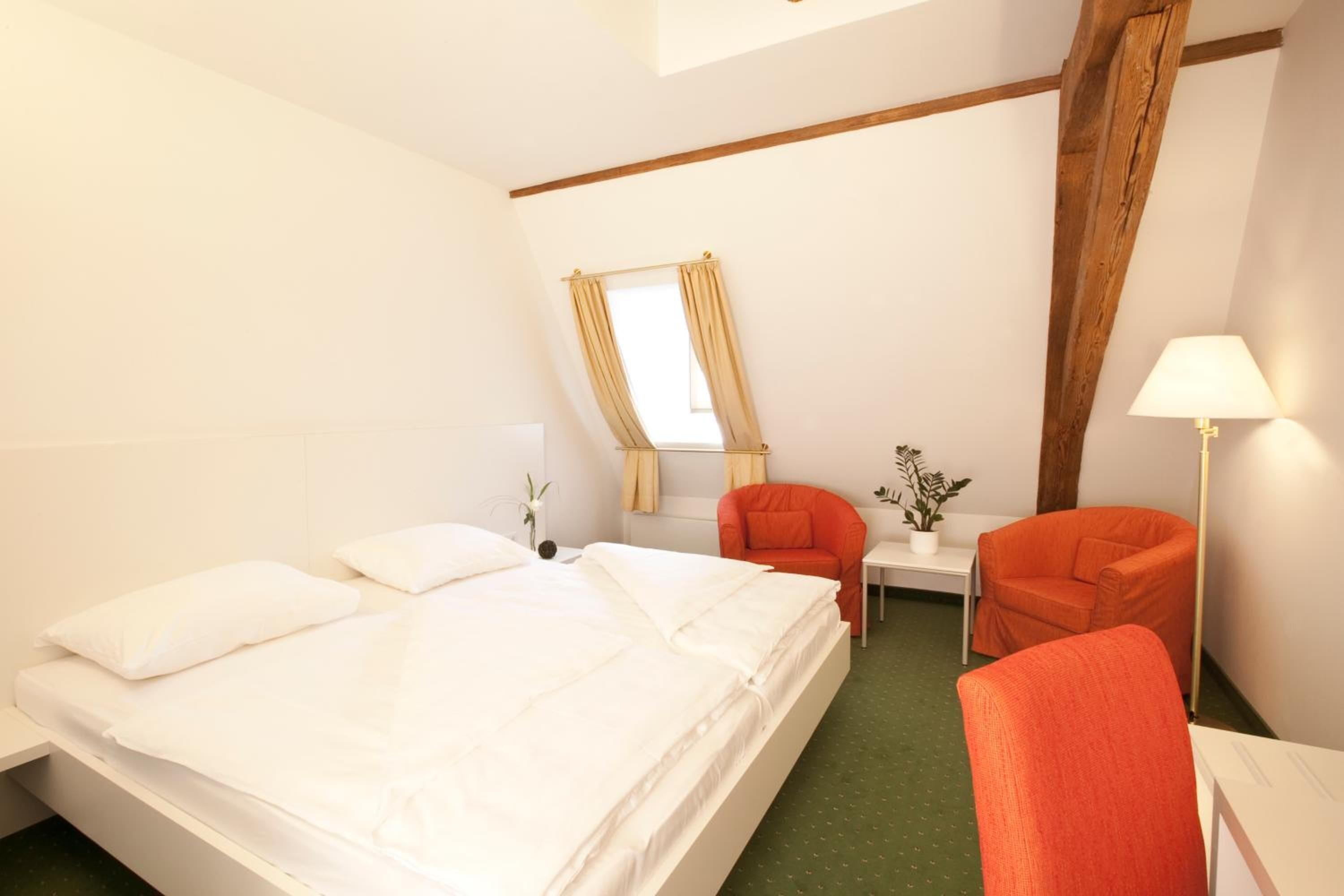 A hotel room with a large bed, a desk, two chairs, a window with curtains, and a lamp.