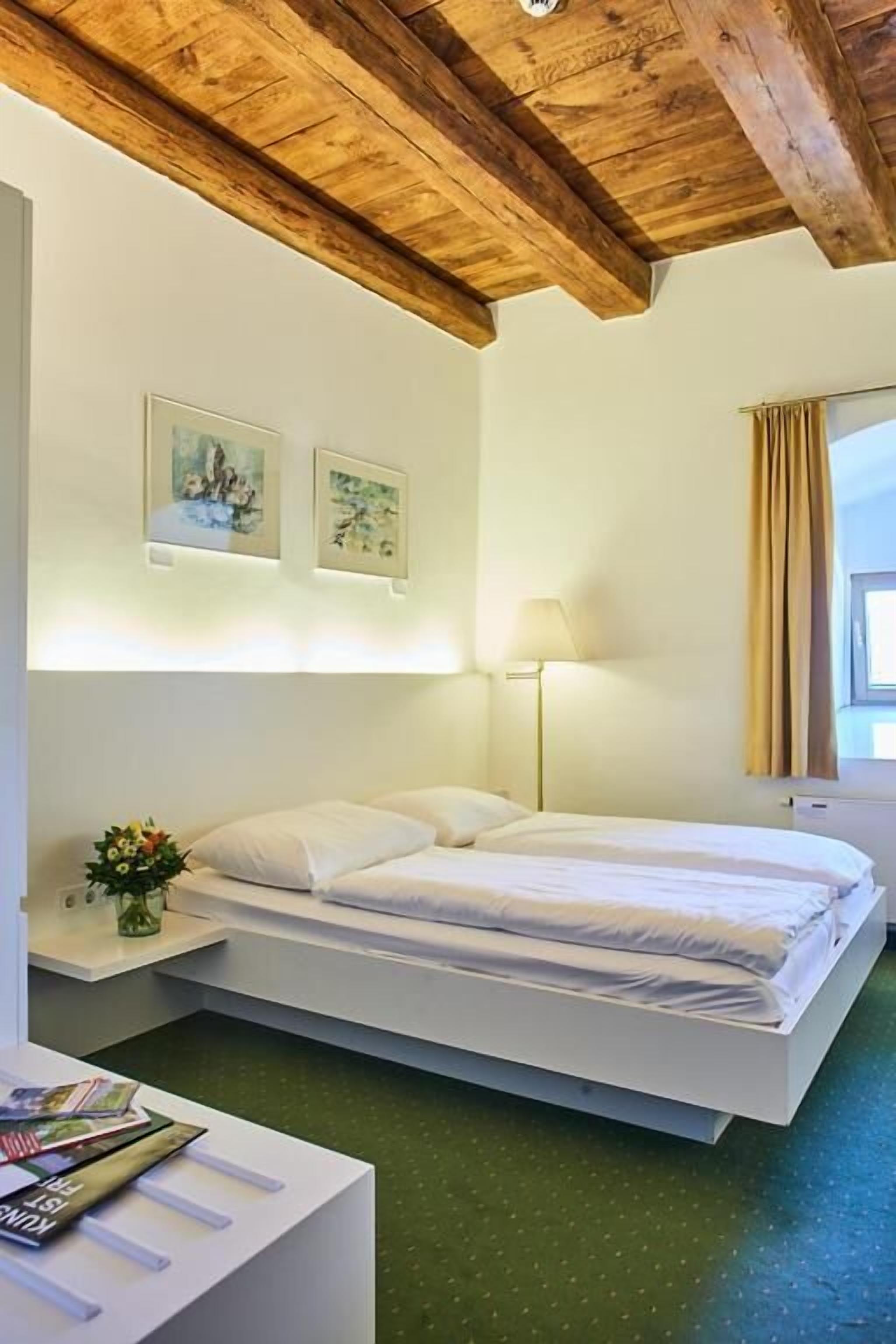 A modern hotel room with a wooden ceiling, a bed with white linens, a bedside table with a lamp, and a window with curtains.