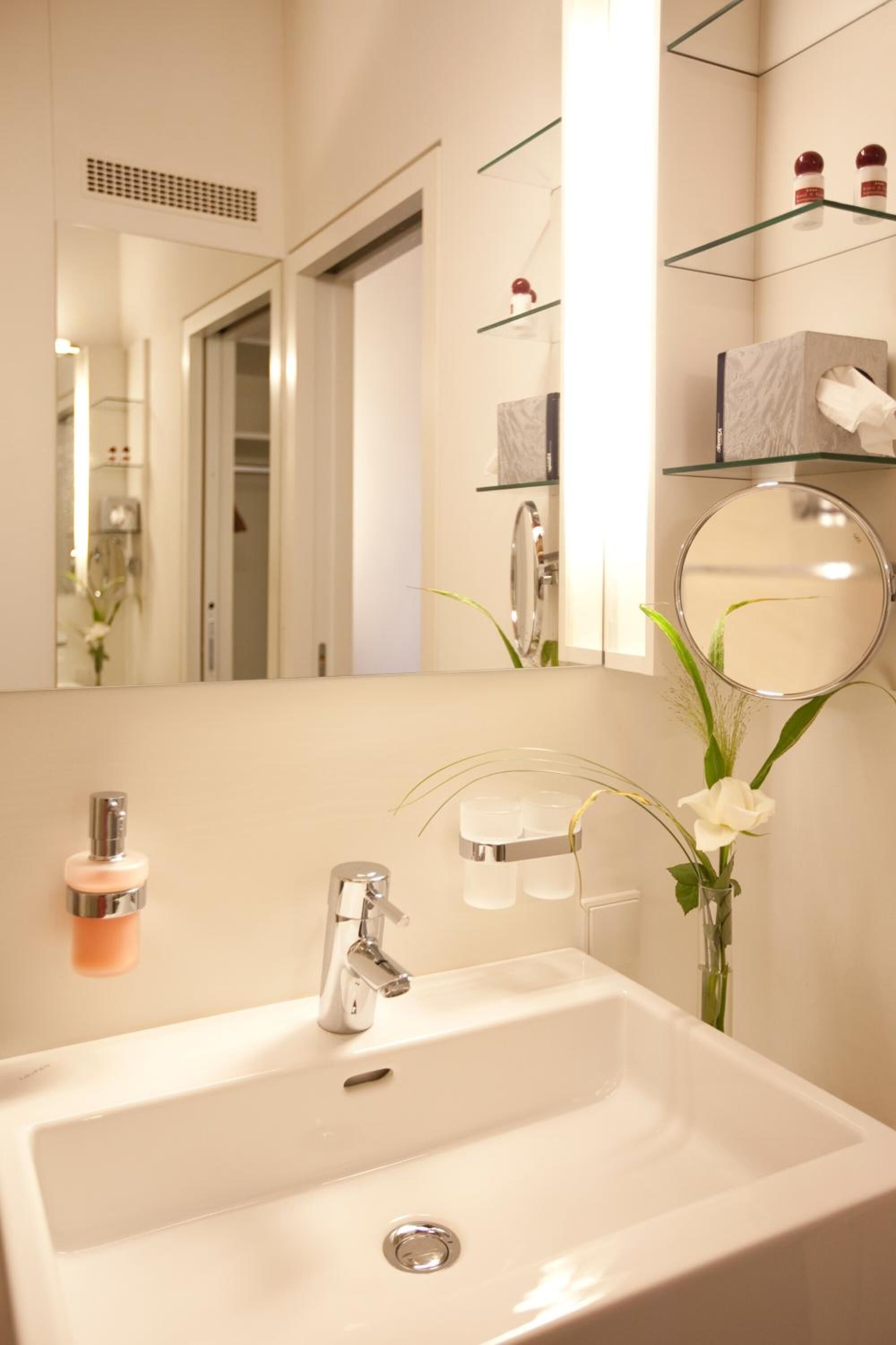 A modern bathroom with a white sink, chrome faucet, and a mirror above it. There are shelves with toiletries and a vase with white flowers on the wall.