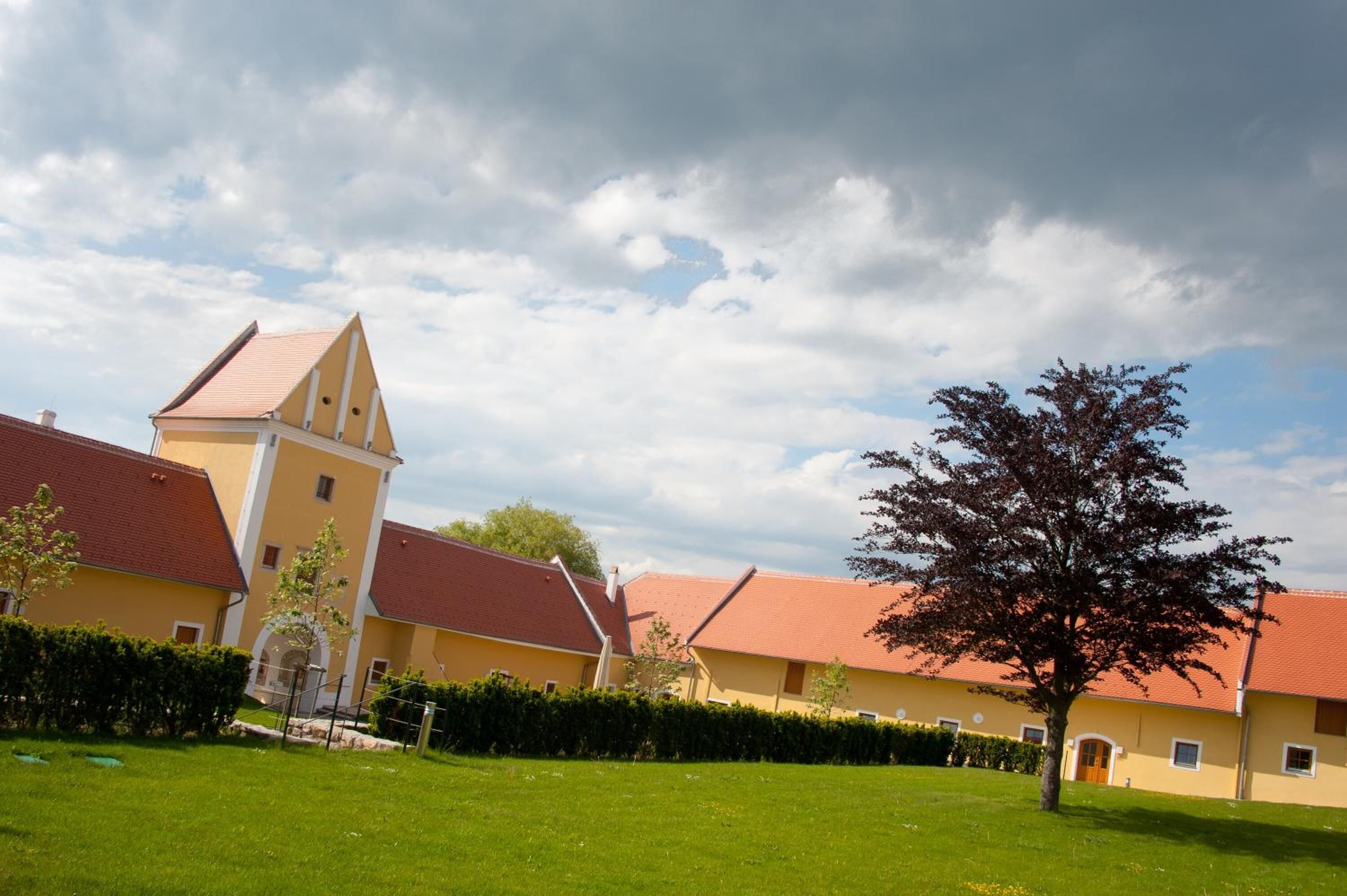 A yellow building with a red roof, surrounded by green grass and a tree.