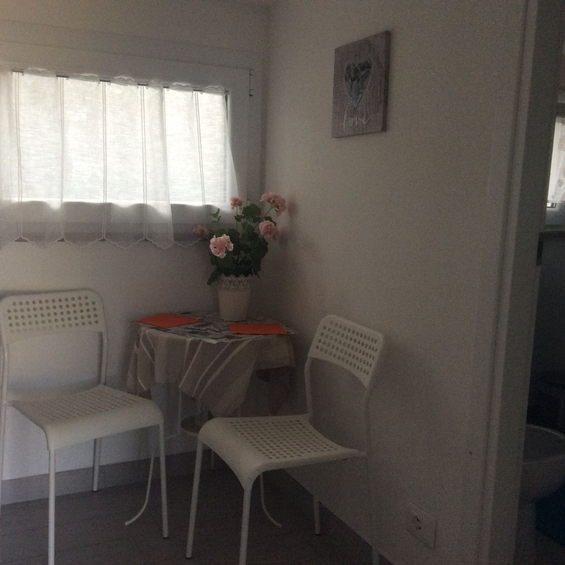 Una habitación pequeña con dos sillas blancas, una mesa con mantel rojo y un jarrón con flores rosadas.