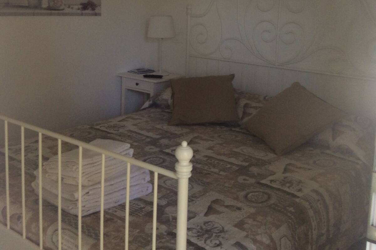 Une chambre à coucher avec un lit, des oreillers, une table de chevet, une lampe, une étagère murale décorée de fleurs et une balustrade blanche.