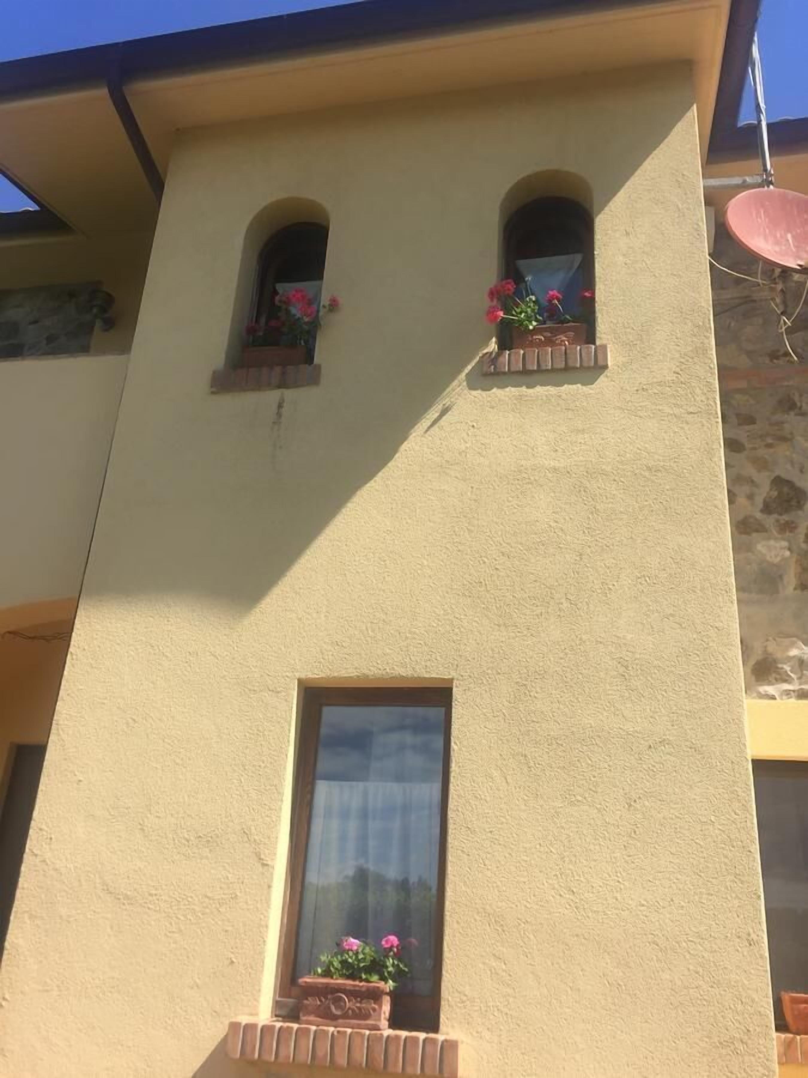 Un bâtiment à l’extérieur de couleur beige, avec un enduit texturé, deux fenêtres en arc surmontées de jardinières, et une grande fenêtre rectangulaire en dessous.