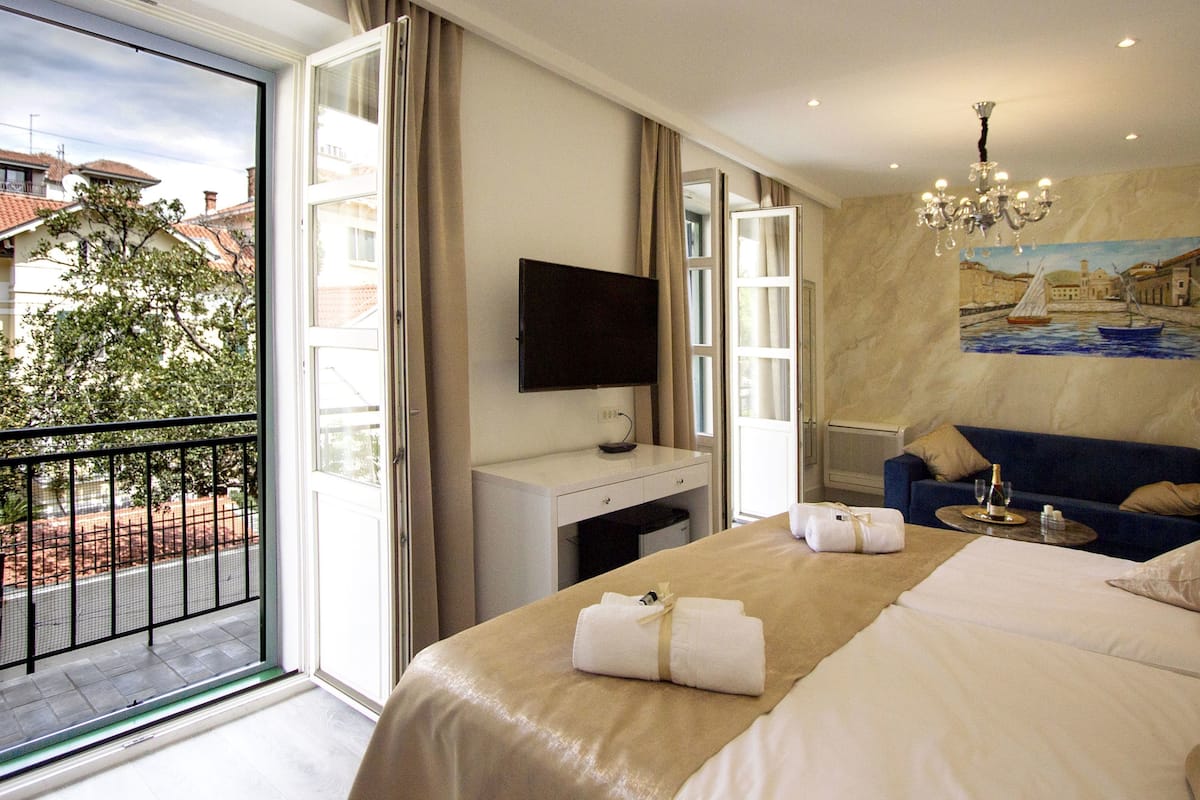 A hotel room with a bed, a desk, a chair, a TV, a painting, and a balcony with a view of buildings and trees.
