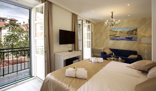 Ein Hotelzimmer mit Bett, Schreibtisch, Sessel, Fernseher, einem Gemälde und einem Balkon mit Blick auf Gebäude und Bäume.