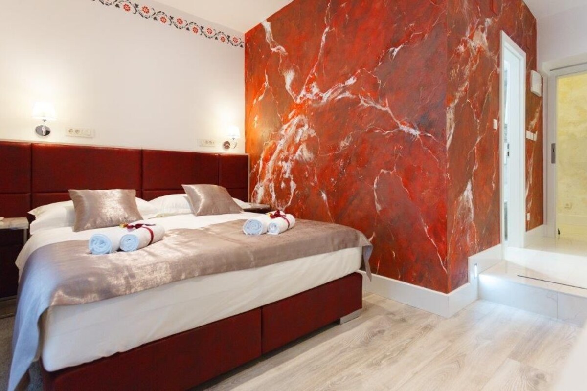A modern hotel room with a large bed, marble wall, and a view of the city.