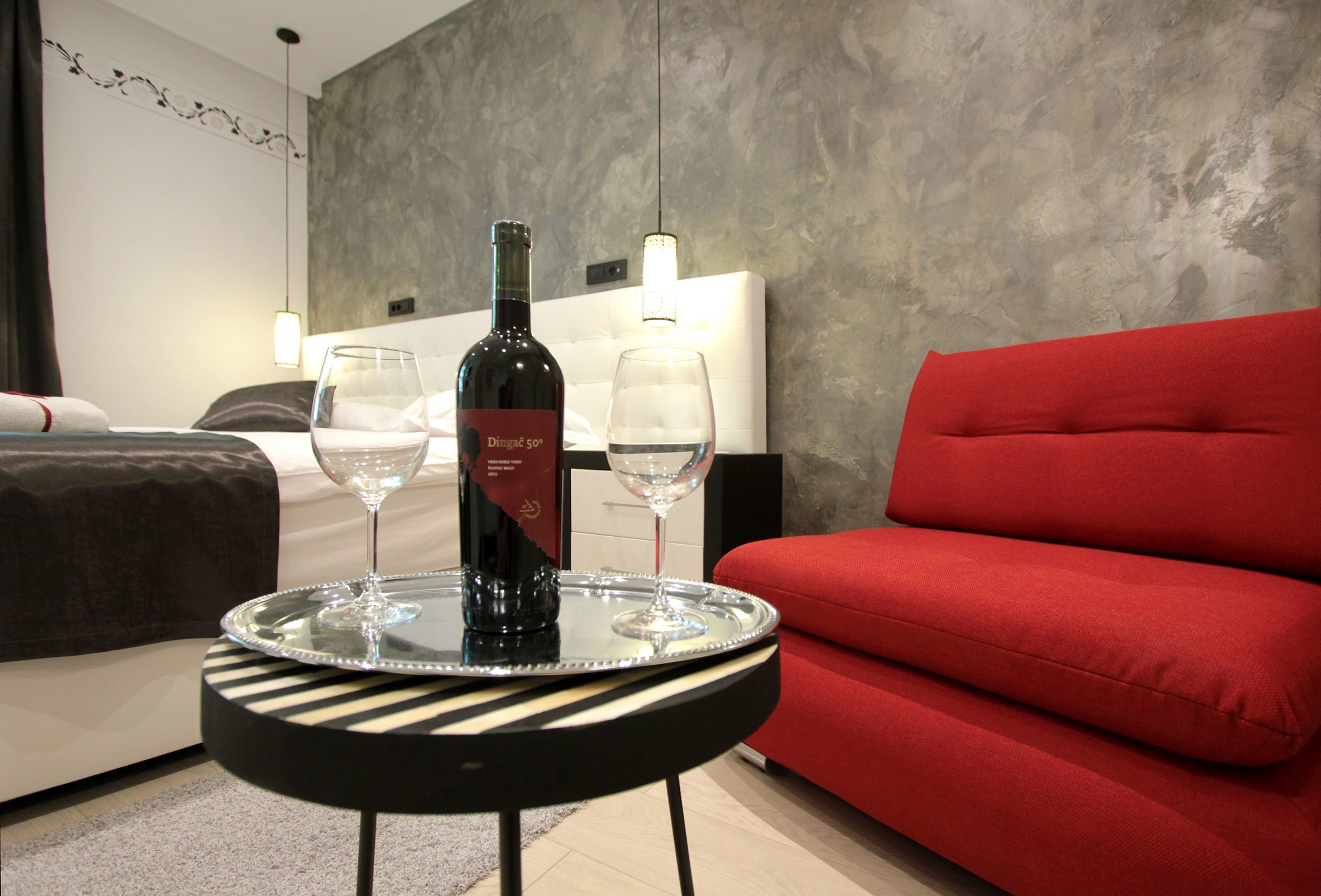 Une chambre d’hôtel avec un lit, un canapé rouge et une petite table ronde sur laquelle se trouve un plateau avec une bouteille de vin et deux verres.