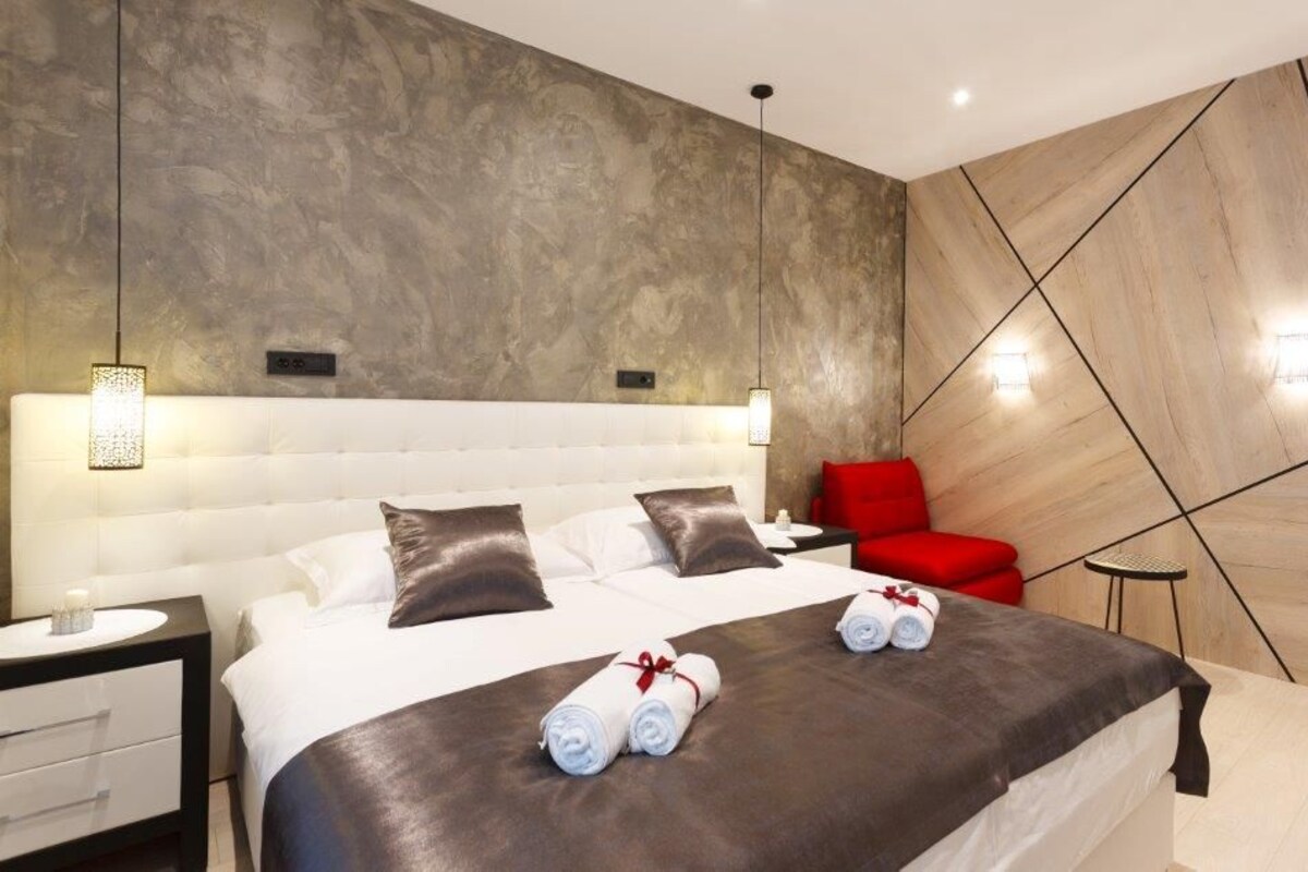 Une chambre d’hôtel moderne avec un grand lit, une table de chevet, un fauteuil rouge et un mur orné de motifs géométriques.