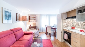Un appartement moderne avec un canapé rouge, une table basse en verre, un coin repas avec une table et des chaises, une cuisine avec des meubles en bois et un crédence carrelée, et une grande fenêtre avec des rideaux légers.