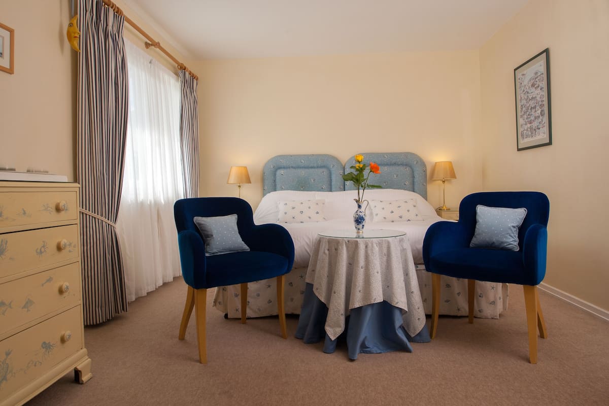 A bedroom with a bed, two chairs, a dresser, a table with a vase, and curtains.