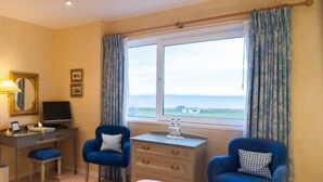 A hotel room with a bed, two blue armchairs, a desk with a computer, a small table with a vase, and a window with a view of the sea. - Sunny Brae (Nairn)