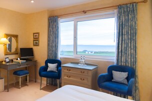 Habitación de hotel con cama, dos sillones azules, un escritorio con computadora, una mesita con un florero y una ventana con vista al mar.