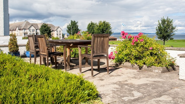 A patio with a wooden table and chairs, surrounded by lush greenery and pink flowers. - Sunny Brae (Nairn)