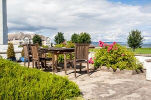Un patio con una mesa y sillas de madera, rodeado de exuberante follaje y flores rosadas.