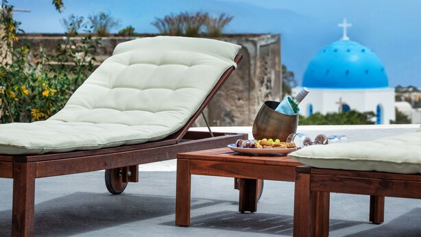 A lounge chair with a cushion, a small table with a plate of food and a drink, and a view of a blue-domed building in the background. - Casa d'Argento (Santorini)