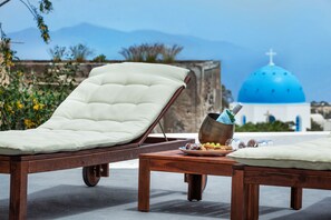 A lounge chair with a cushion, a small table with a plate of food and a drink, and a view of a blue-domed building in the background. - Casa d'Argento (Santorini)