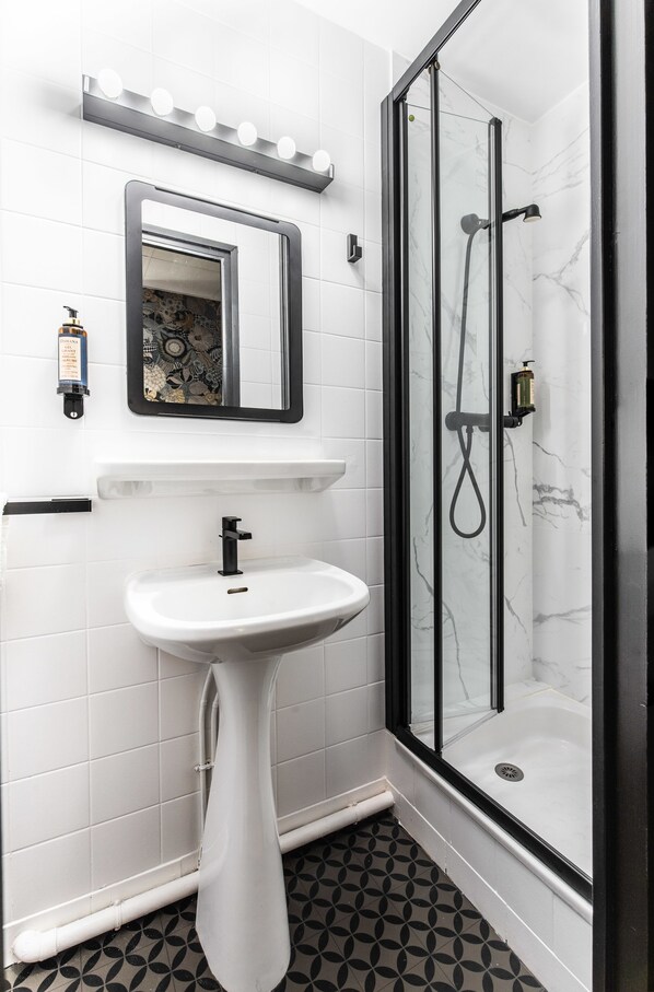 A bathroom with a white pedestal sink, a mirror with lights, a glass-enclosed shower, and patterned floor tiles. - Hôtel Lanjuinais (Rennes)