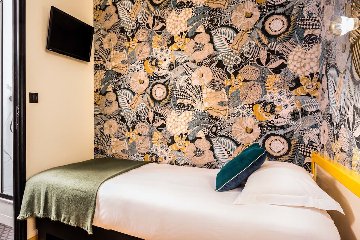 A bedroom with a patterned wallpaper, a bed with a green pillow, a bedside table, a mirror, and a TV mounted on the wall.