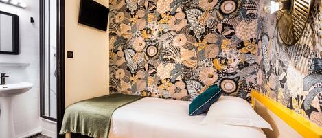 A bedroom with a patterned wallpaper, a bed with a green pillow, a bedside table, a mirror, and a TV mounted on the wall.