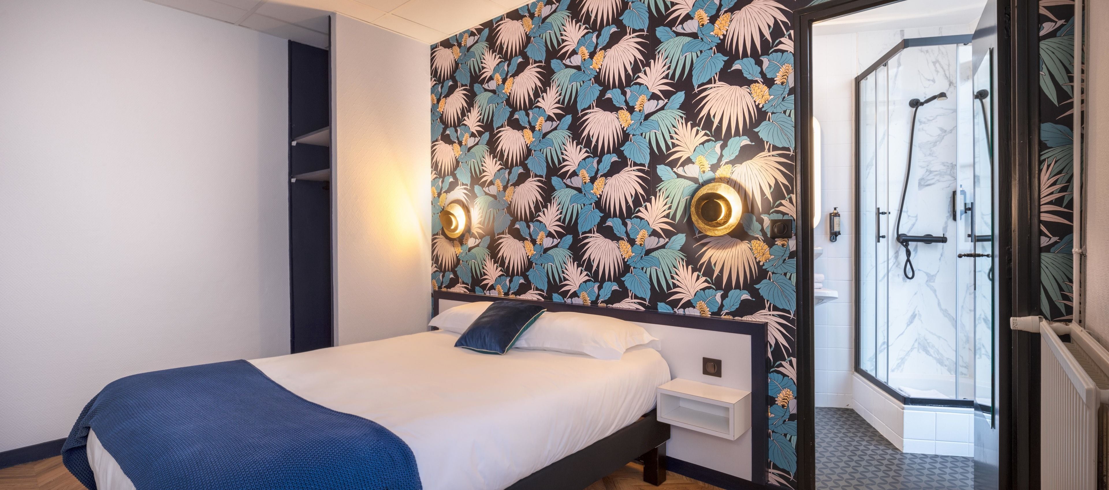A hotel room with a bed, a nightstand, a wall with a tropical leaf pattern, and a shower area with a glass door.