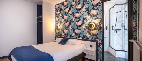 Habitación de hotel con cama, mesita de noche, pared con estampado de hojas tropicales y zona de ducha con puerta de vidrio.