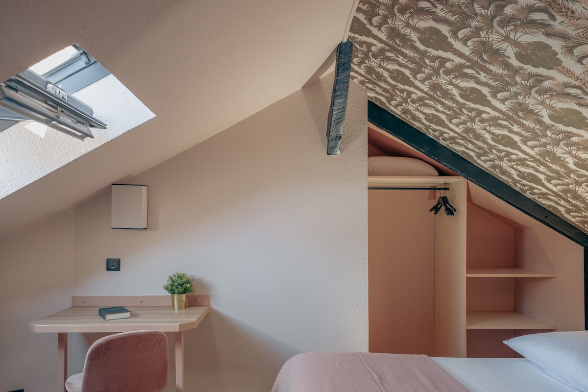 A bedroom with a sloped ceiling, a desk with a chair, a bed, and a small plant.