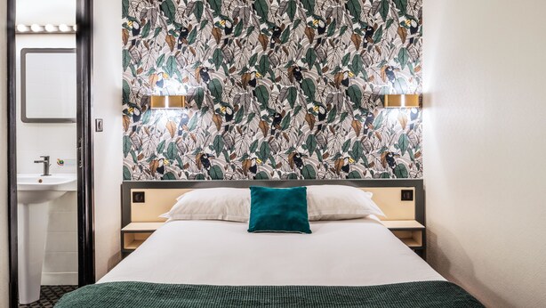 Habitación de hotel con cama, mesita de noche, espejo en la pared y baño a la vista a través de una puerta abierta.