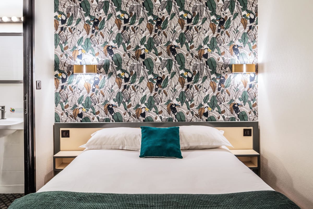 A hotel room with a bed, a nightstand, a wall-mounted mirror, and a bathroom visible through an open door.