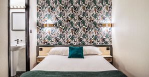 Habitación de hotel con cama, mesita de noche, espejo en la pared y baño a la vista a través de una puerta abierta.