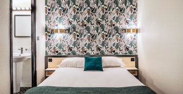 Une chambre d’hôtel avec un lit, une table de chevet, un miroir fixé au mur et une salle de bain visible à travers une porte ouverte.