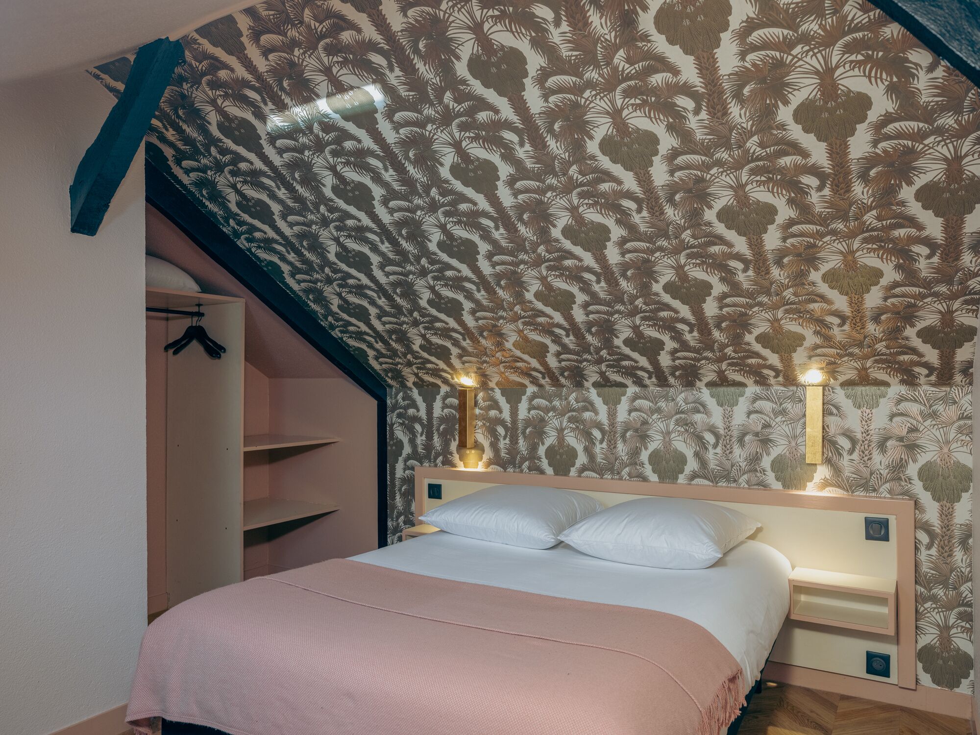 A bedroom with a sloped ceiling, a bed with white bedding, two bedside lamps, and a wardrobe with a hanging rack.