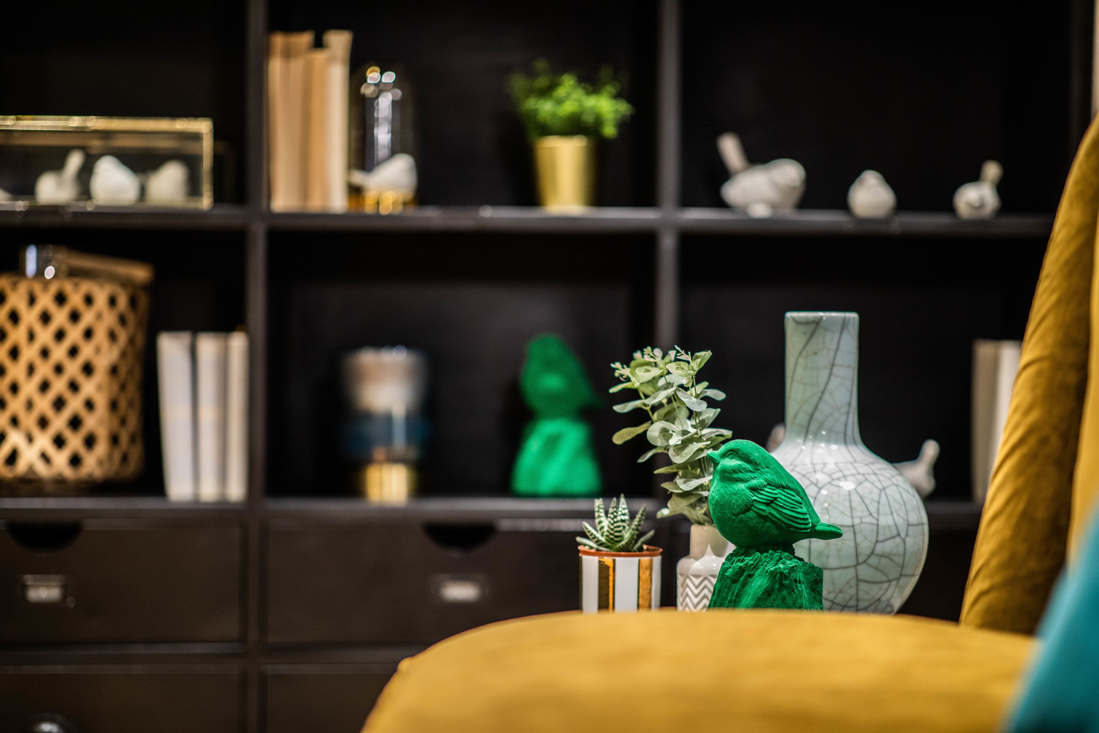 A room with a yellow sofa, a white vase with a green plant, and a shelf with decorative items.