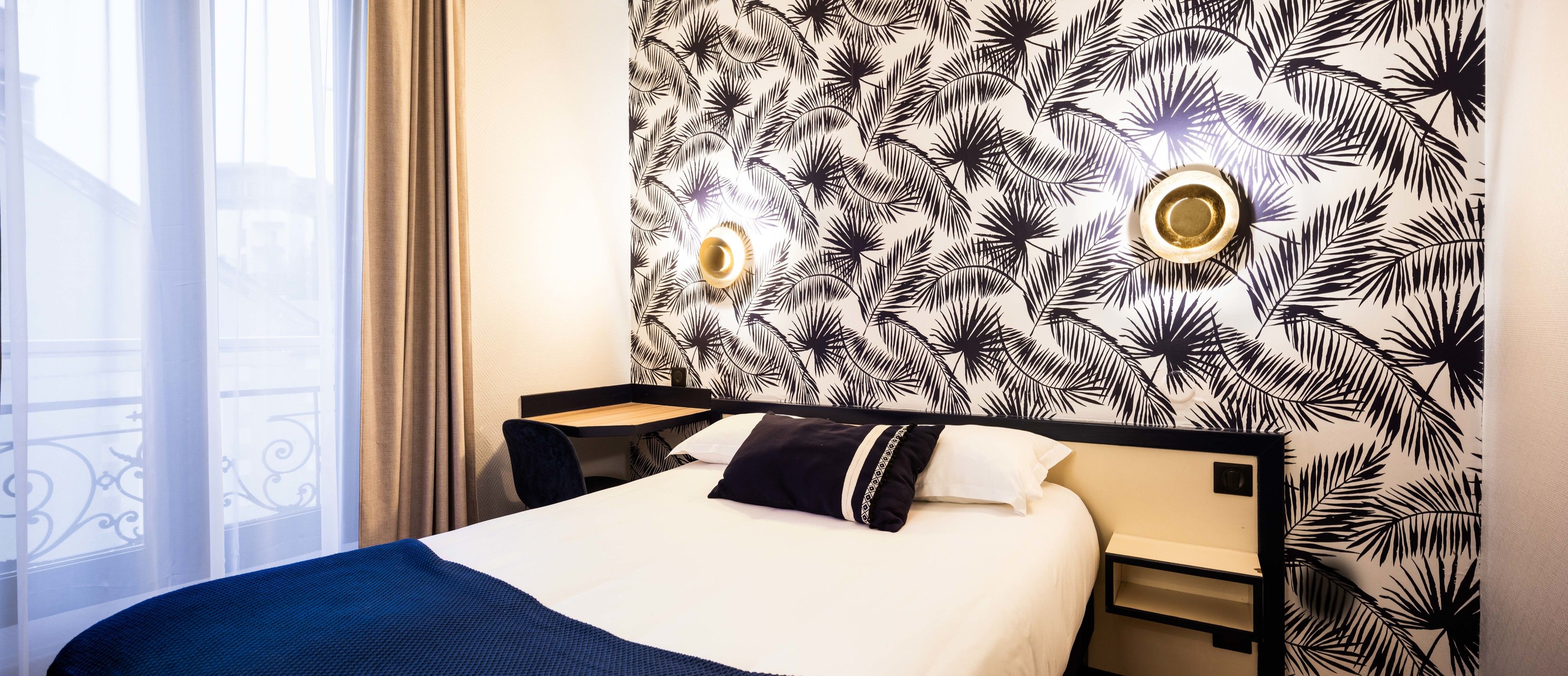 Habitación de hotel con cama, mesita de noche, pared con diseño de hojas y ventana con vista a un balcón.