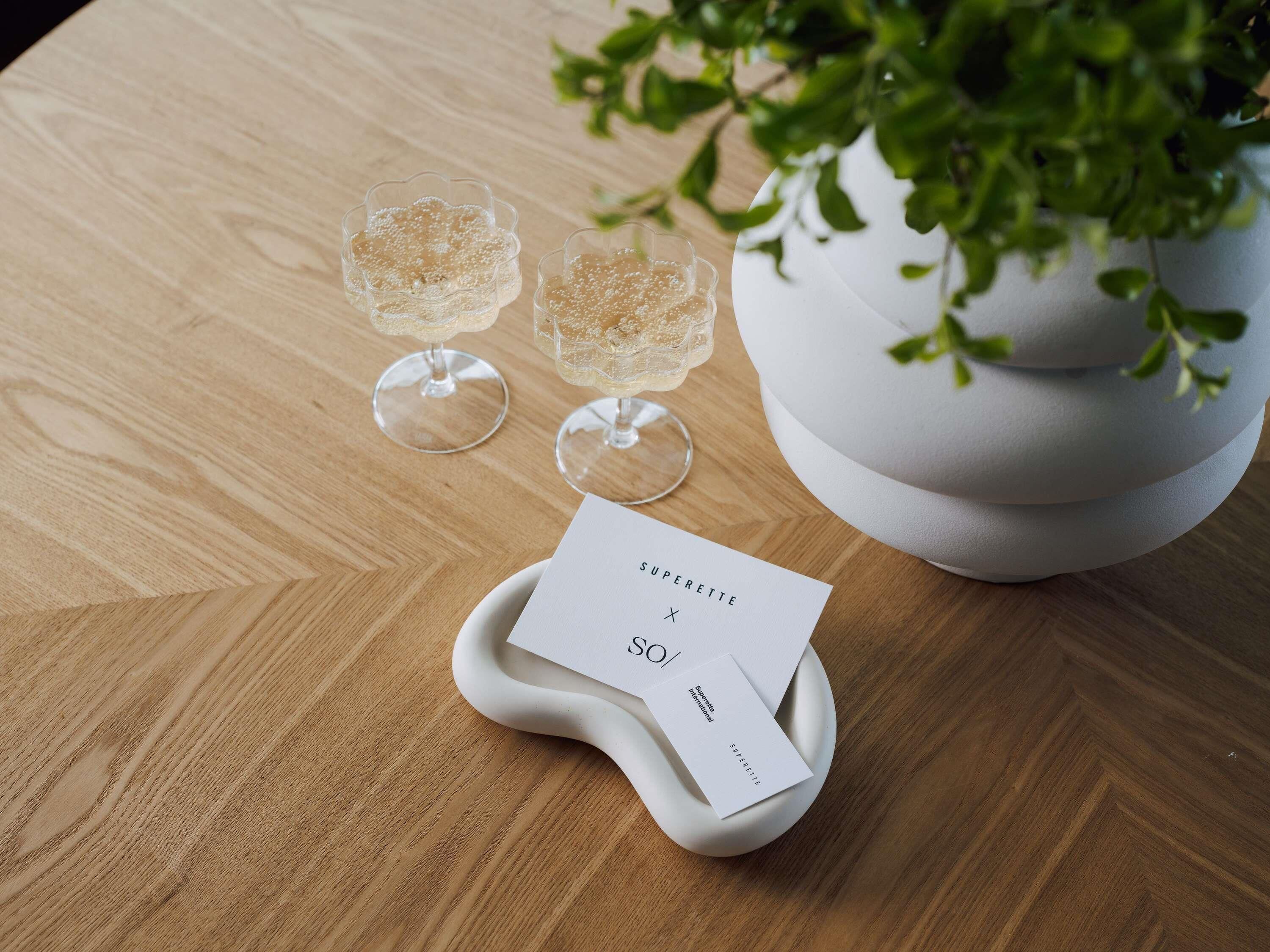 A wooden table with two glasses of a bubbly beverage, a white vase with green leaves, and a card labeled 'SUPERETTE.'