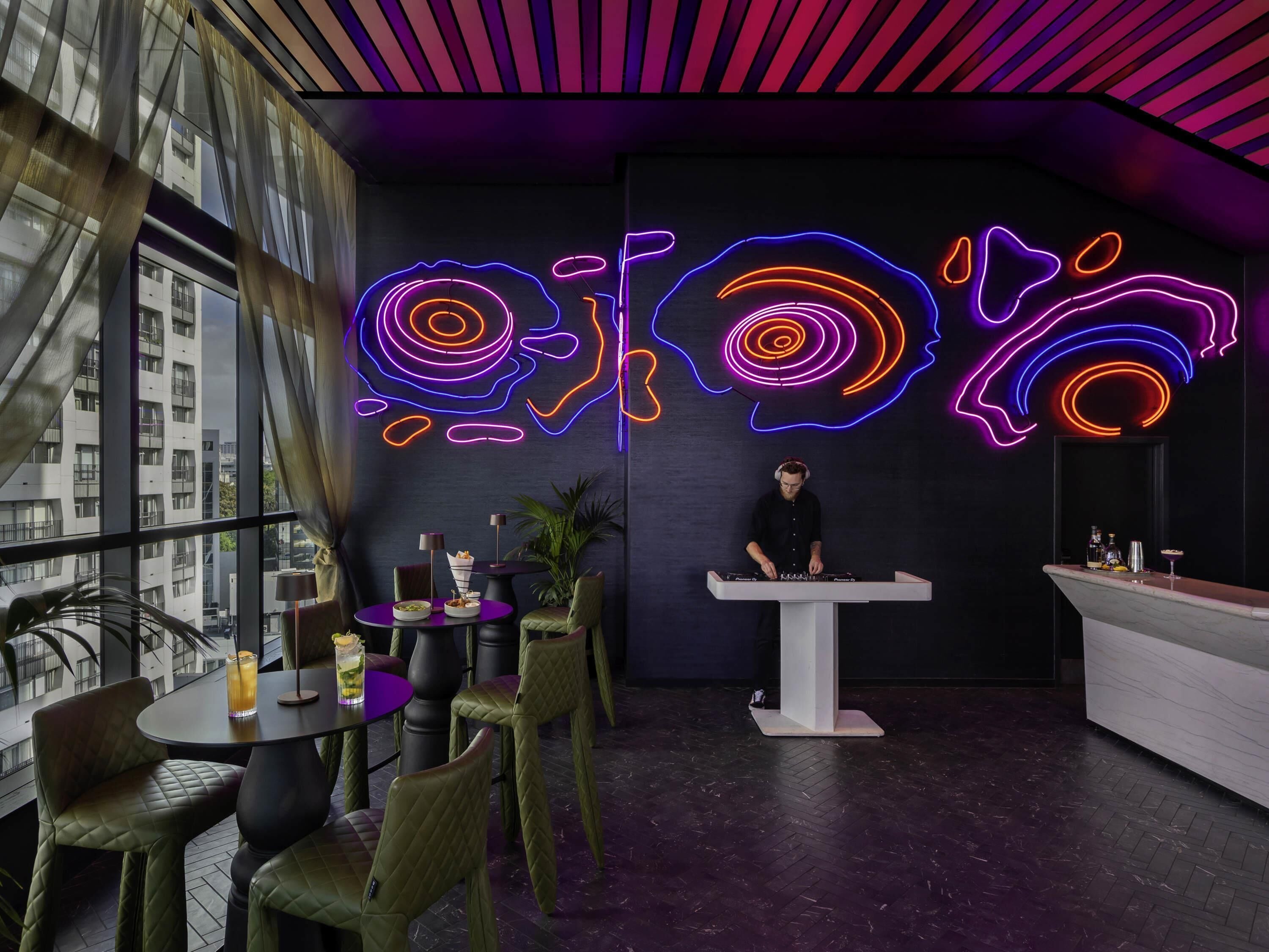 A modern bar area with neon art, a DJ booth, and a view of buildings through large windows.