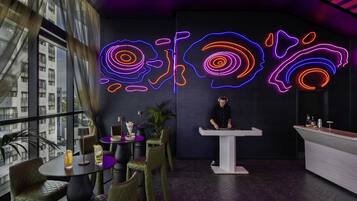 A modern bar area with neon art, a DJ booth, and a view of buildings through large windows.
