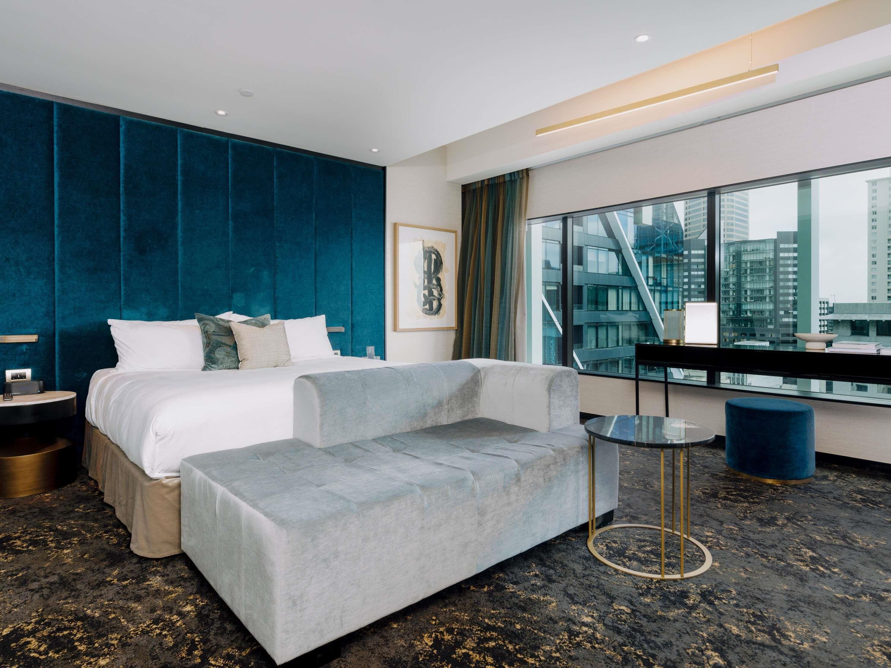 A modern hotel room with a large bed, a grey sofa, a small table, and a view of the cityscape through large windows.