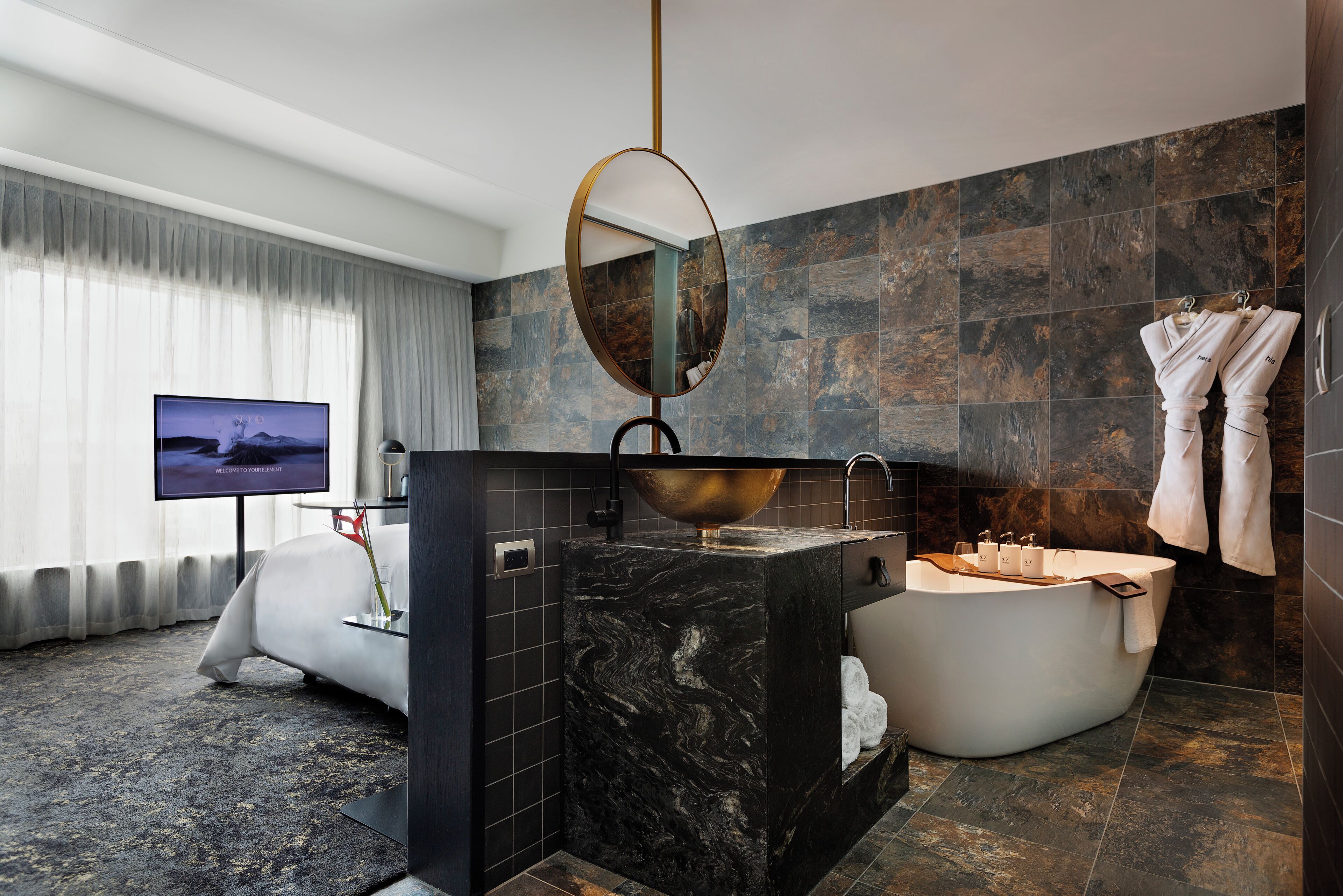 A modern bathroom with a large bathtub, a round mirror, and a television on a stand.