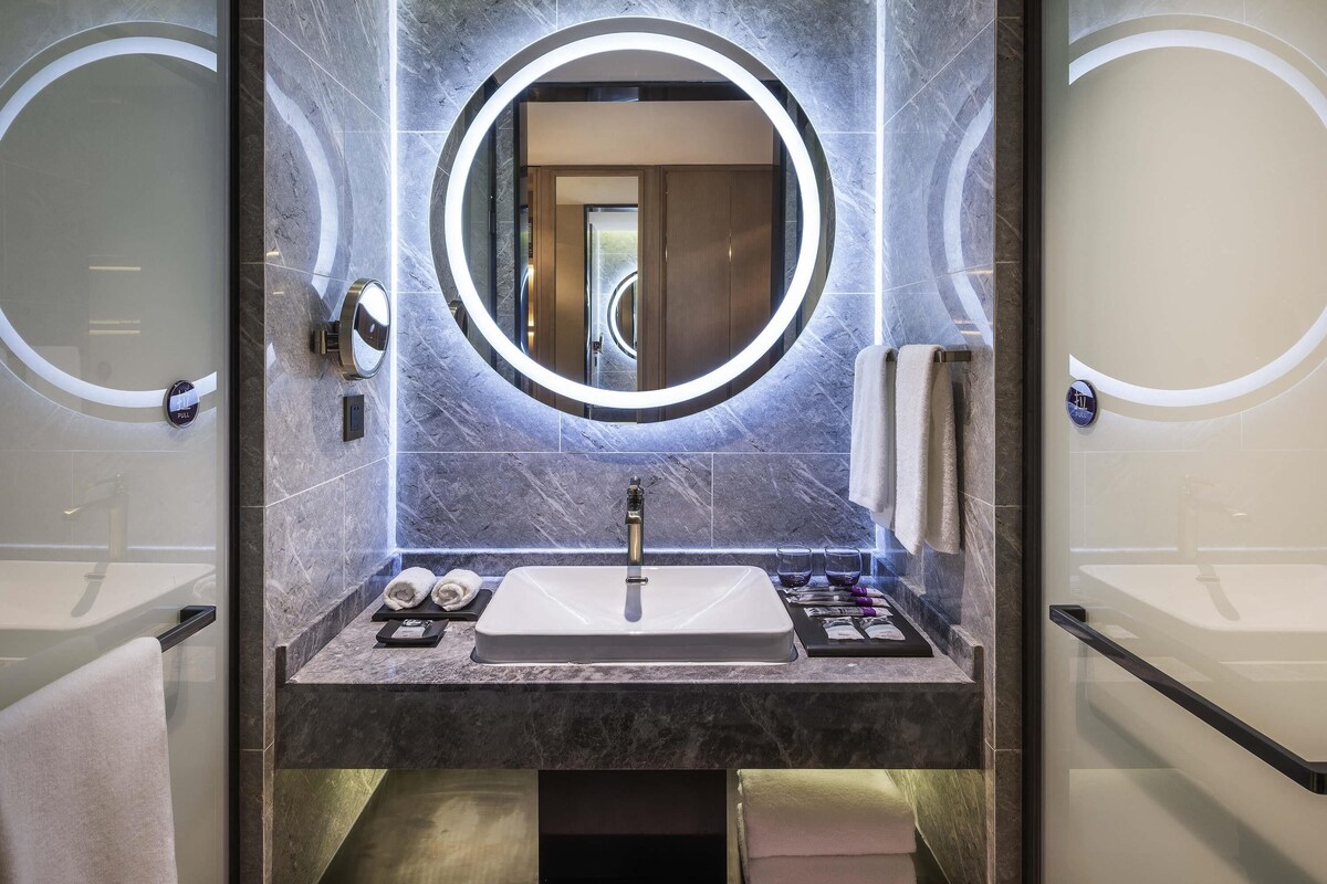 A modern bathroom with a central sink, a mirror with a circular light, and two towel racks.